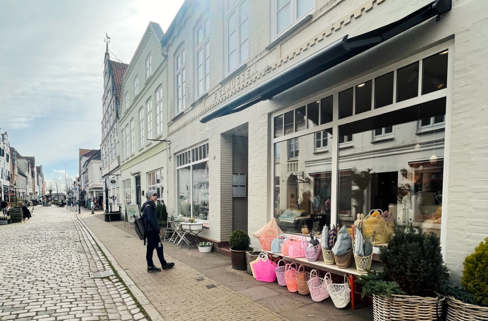 Sonnenbeschienene Straße mit historischen Gebäuden; Mann geht am 'Nest Concept Store' vorbei, vor dem bunte Taschen ausgestellt sind.